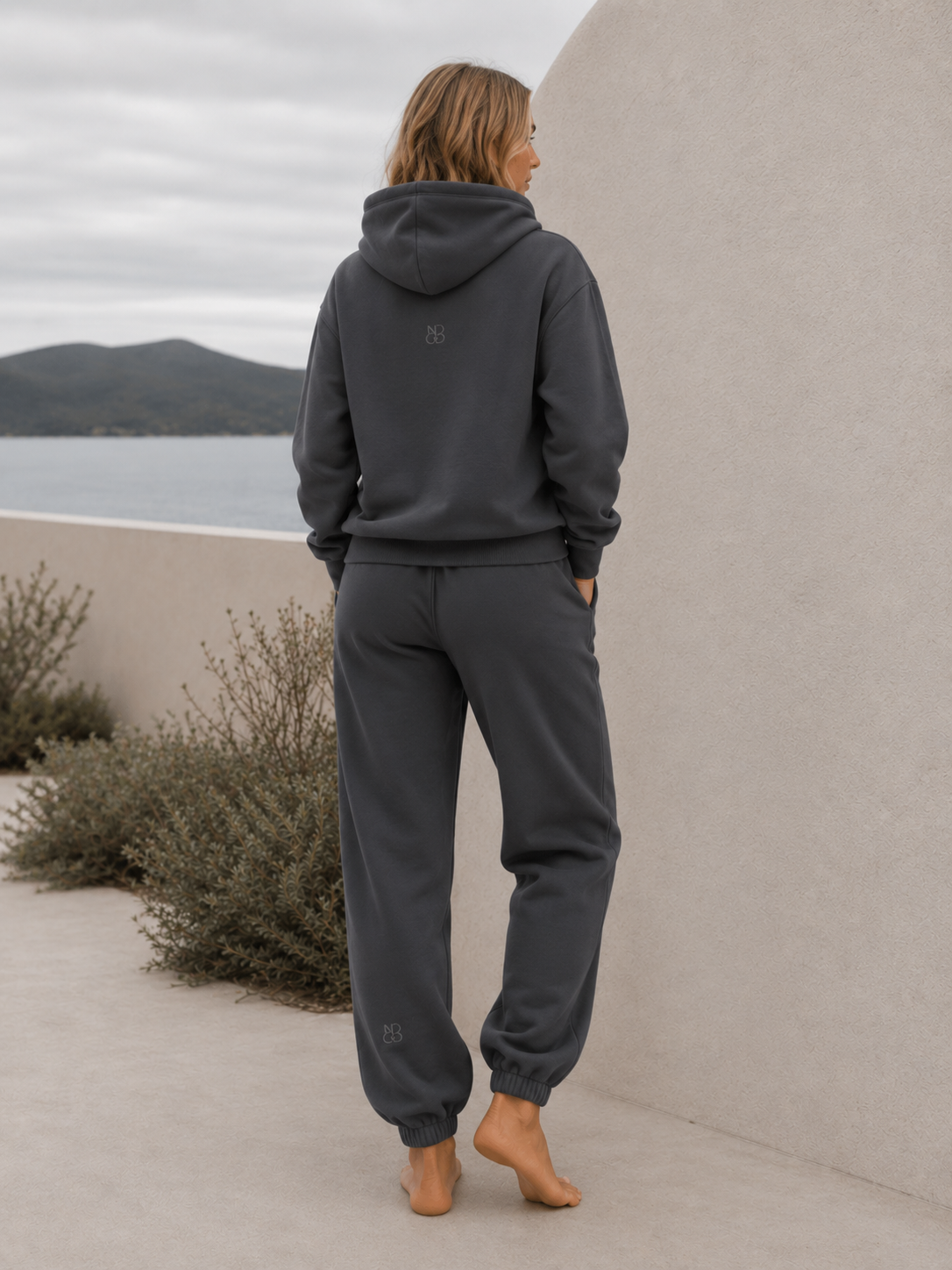 Woman wearing the NORTHE Core Classic Sweatpants in Essential Tone Carbon with a matching hoodie, shown from the back in a coastal lifestyle setting.