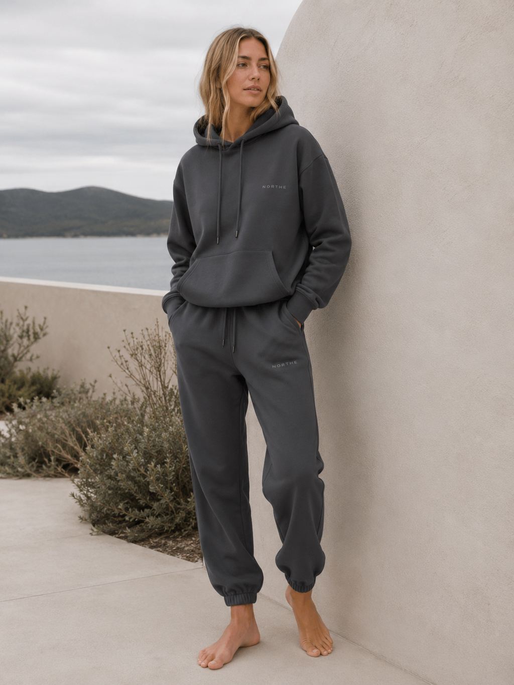 Woman wearing the NORTHE Core Classic Sweatpants in Essential Tone Carbon with a matching hoodie in a coastal lifestyle setting.