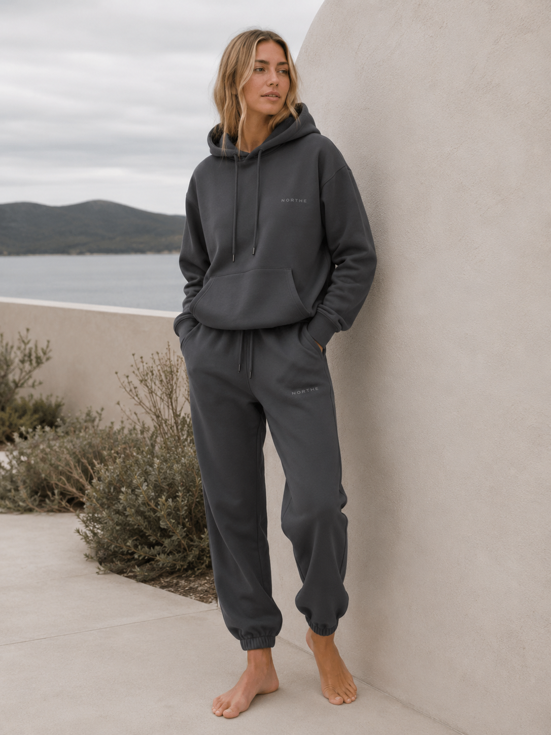 Woman wearing the NORTHE Core Classic Sweatpants in Essential Tone Carbon with a matching hoodie in a coastal lifestyle setting.