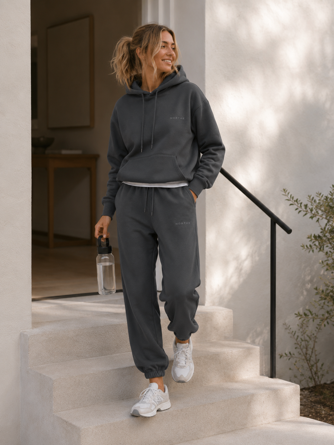 Woman wearing the NORTHE Core Classic Sweatsuit in Essential Tone Carbon, walking down stairs in a relaxed lifestyle setting.