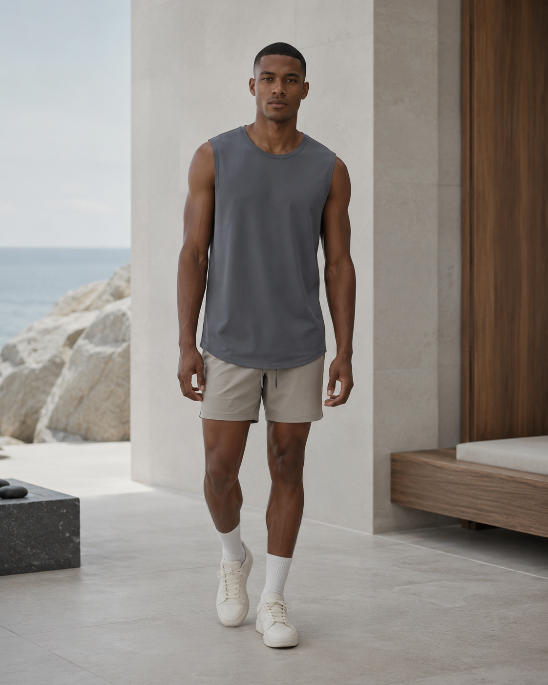 A man modeling a sleeveless crewneck athletic tank top in heather gray, paired with beige shorts, white socks, and white sneakers, standing in a modern outdoor patio setting with a view of the ocean.
