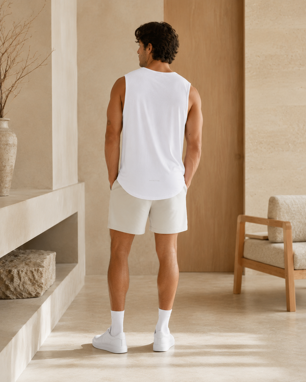 A man viewed from behind wearing a white sleeveless tank top and light beige shorts in a minimalist, warm-toned indoor setting.