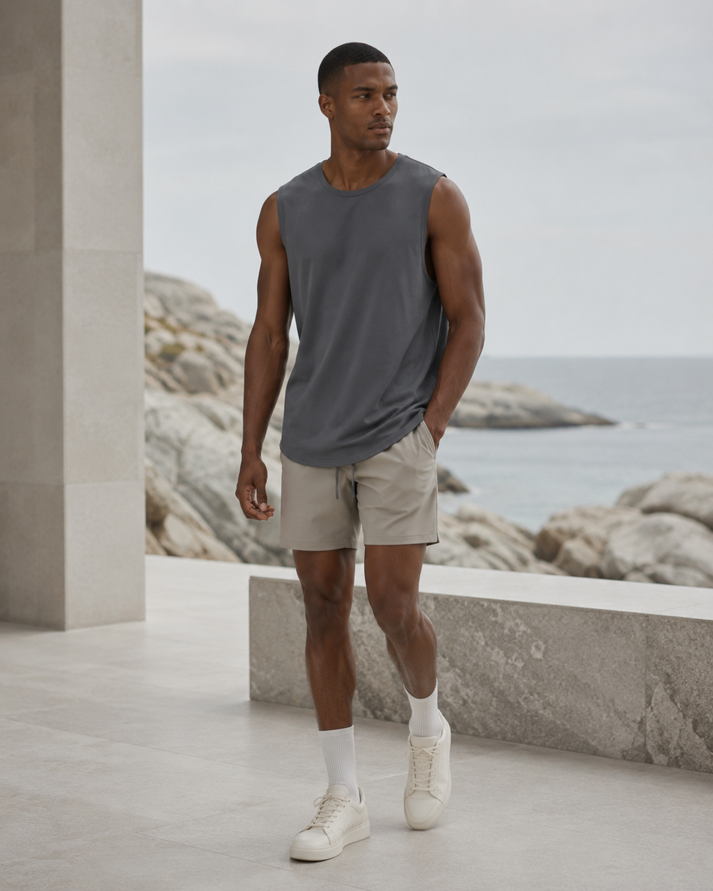 A man wearing a sleeveless dark gray t-shirt, beige shorts, white socks, and white sneakers, standing on a stone terrace with a rocky coastline and ocean in the background.